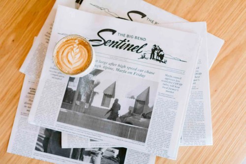 A cup of latte with latte art sits atop a stack of newspapers titled "The Big Bend Sentinel."