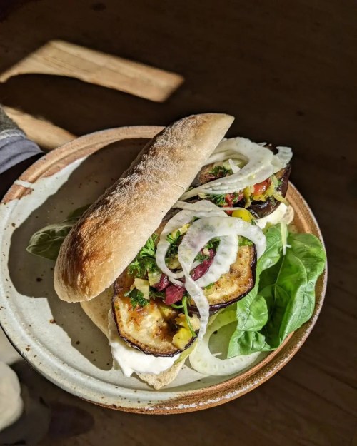 A sandwich with grilled eggplant, greens, onions, and sauce, served on a rustic plate in natural light.