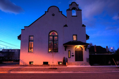 A historic building with stained glass windows, illuminated at dusk, featuring a bell tower and a welcoming entrance.
