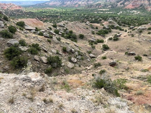 A rocky, arid landscape with scattered shrubs and a winding path, set against a backdrop of distant hills and valleys.