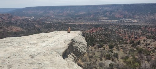 A rocky cliff edge overlooks a vast, rugged landscape with distant hills and valleys under a cloudy sky.