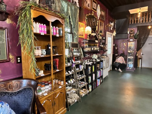 A cozy shop interior featuring shelves of candles, herbs, and various items, with a vintage armchair in the foreground.