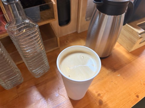 A white cup of coffee sits on a wooden table, surrounded by glass bottles and a metal coffee carafe.