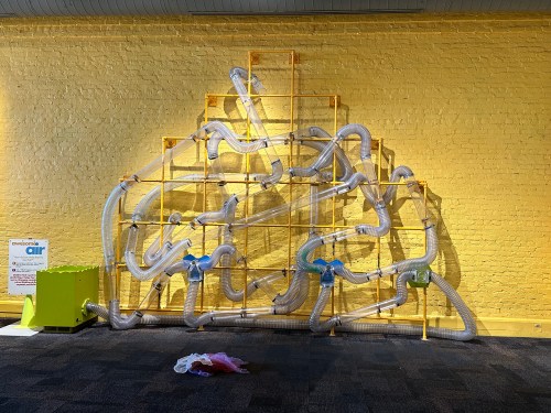 A colorful wall installation with clear tubes arranged in a playful design against a yellow background.