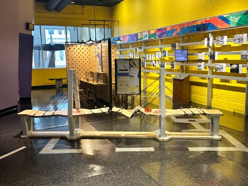 A wooden bridge model with cables, displayed in a brightly colored exhibit space with informational panels.