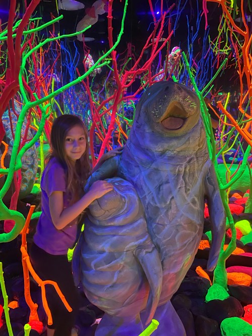 A girl poses with a colorful, oversized manatee sculpture in a vibrant, neon-lit environment.