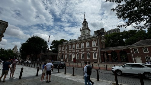A historic building with a clock tower, surrounded by trees and people walking on a cloudy day.