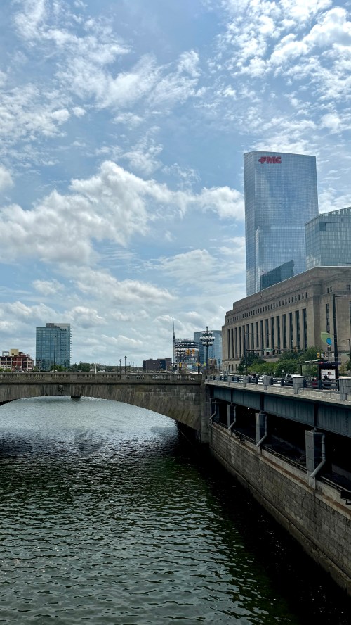 A cityscape featuring a river, a bridge, and modern buildings under a partly cloudy sky.