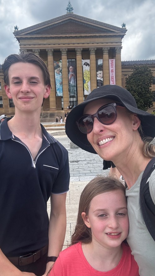 A woman and two children pose for a selfie in front of a large museum with colorful banners.