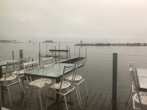 Foggy waterfront view with empty tables and chairs on a wooden deck, overlooking calm water and distant boats.