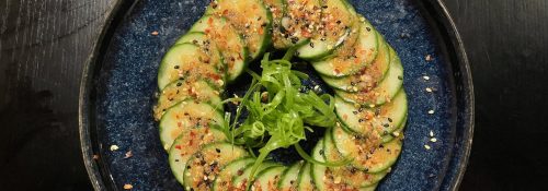 A circular plate of sliced cucumbers topped with sesame seeds, green onions, and spices on a dark background.
