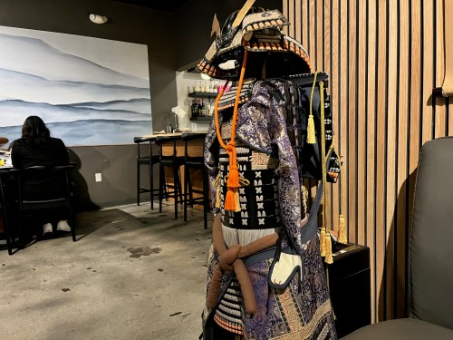 A decorative samurai armor displayed in a modern setting, with wooden paneling and a blurred figure in the background.