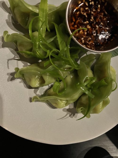 A plate of green dumplings garnished with green onions, accompanied by a small bowl of dipping sauce.