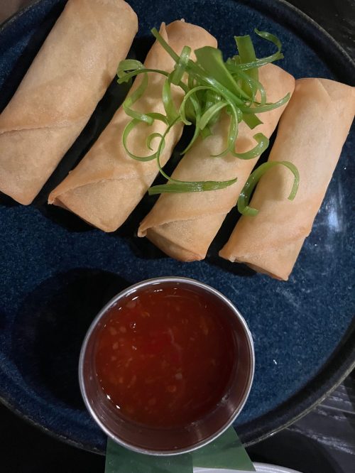 Four crispy spring rolls on a blue plate, garnished with green onions, accompanied by a small bowl of dipping sauce.