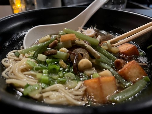 A close-up of a bowl of noodle soup topped with green onions, mushrooms, and tofu, with chopsticks resting on the side.