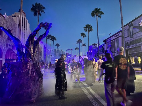 A spooky street scene at dusk with costumed figures, palm trees, and fog, creating a festive Halloween atmosphere.