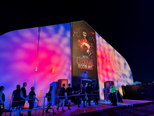 A crowd lines up outside a brightly lit building featuring a large poster for "Eternal Bloodlines" at night.