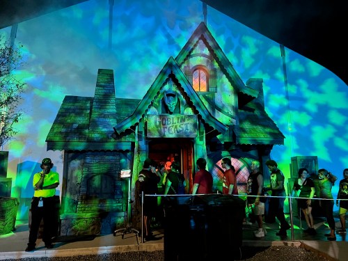 A spooky house facade with colorful projections, people lined up outside, and a security guard nearby.