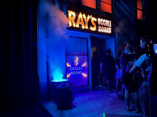 A dimly lit storefront with "Ray's Occult Books" sign, smoke effects, and a line of people waiting to enter.