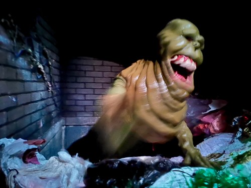 A grotesque green creature with a wide mouth emerges from a pile of trash in a dark, eerie setting.