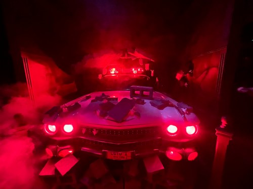 A vintage car surrounded by red lighting, smoke, and scattered debris, creating a dramatic and eerie atmosphere.