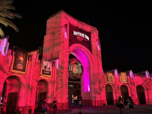 Universal Studios entrance illuminated in red for Halloween Horror Nights, with palm trees and visitors in the foreground.