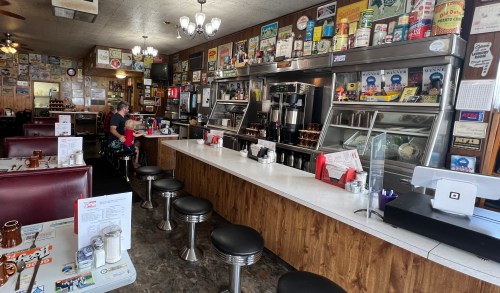 A retro diner interior with a long counter, stools, and vintage decor, featuring a customer and a server.