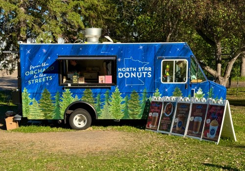 A colorful food truck named "North Star Donuts" parked in a park, surrounded by trees and displaying menu signs.