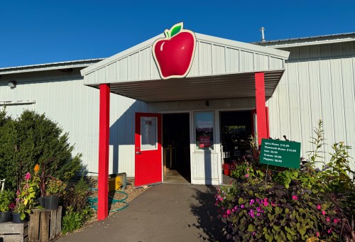 Entrance of a market with a large apple logo, red door, and colorful flowers in front. Clear blue sky above.