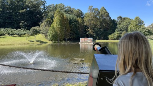 A child watches a pond with a fountain and trees, while a colorful mural is visible on a wall in the background.