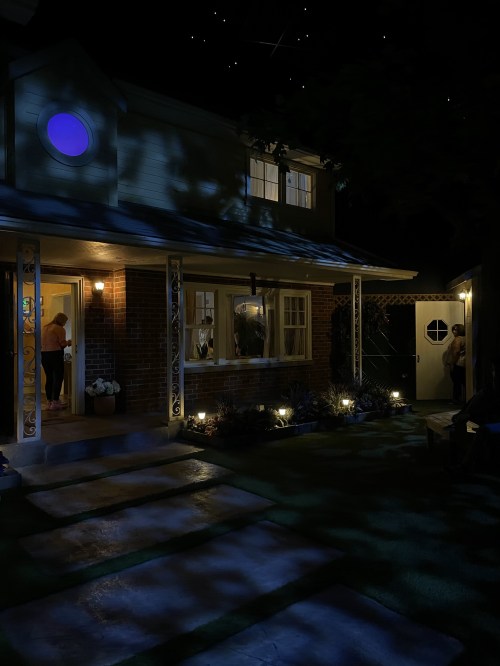A dimly lit house at night, featuring a blue circular window, illuminated pathways, and people visible inside.