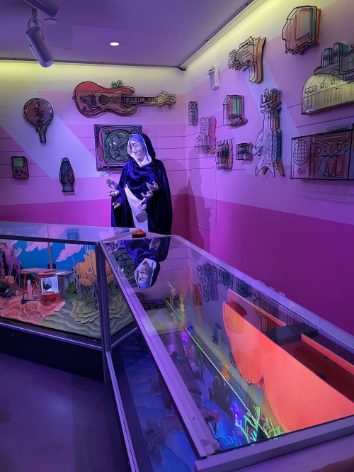 A colorful exhibit featuring a statue of a figure in a robe, surrounded by artistic wall displays and illuminated glass cases.