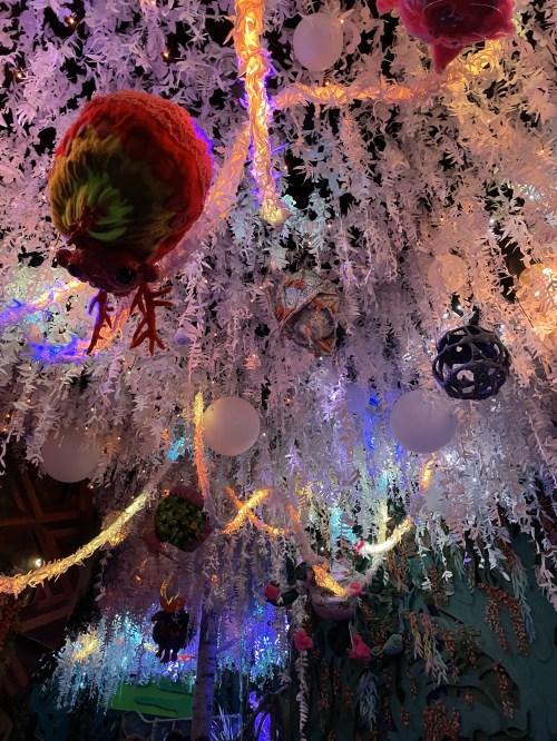 Colorful decorations hang from a ceiling covered in white foliage, illuminated by vibrant lights and unique ornaments.