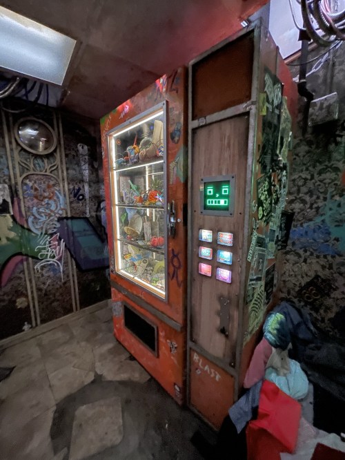 A vintage vending machine with a glass display, covered in stickers, in a graffiti-decorated room.