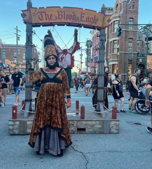 A performer in a dark, ornate costume stands near a medieval-themed display with a sign reading "The Blood Eagle."