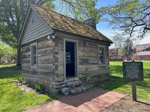 A small log cabin with a mossy roof, surrounded by greenery, featuring a sign that reads "Post Office."
