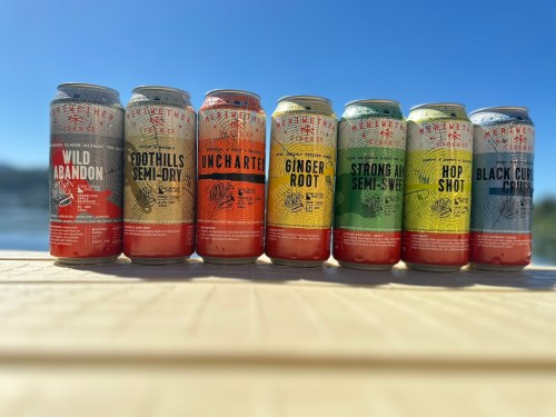 A row of colorful craft beer cans on a table, with a scenic outdoor background.