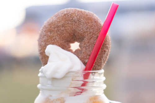 A close-up of a milkshake topped with whipped cream and a cinnamon sugar donut, with a pink straw inserted.