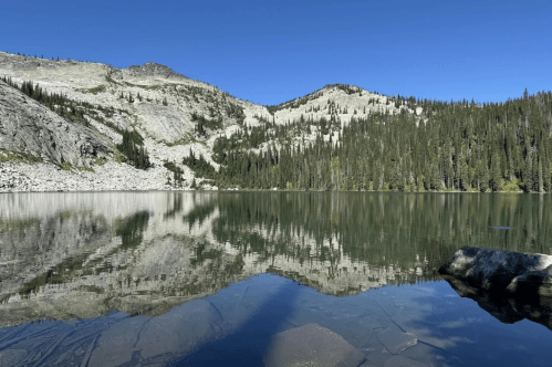 A serene lake reflecting mountains and trees under a clear blue sky, with a rocky shore in the foreground.