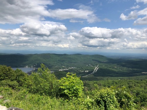 A panoramic view of lush green mountains under a partly cloudy sky, with a winding road and a lake in the distance.