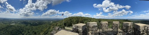 Panoramic view from a rocky overlook, featuring lush green hills, a blue sky with clouds, and an American flag.