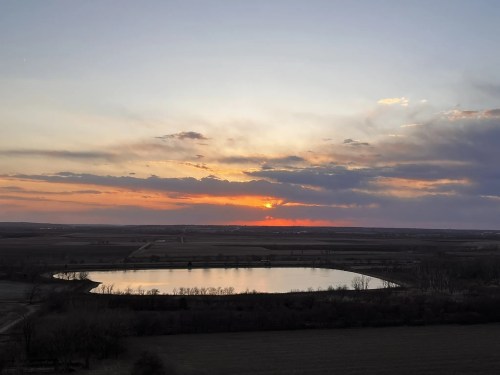 Sunset over a calm lake, with colorful clouds reflecting on the water and a vast landscape in the background.
