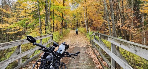A bike rests on a wooden bridge overlooking a colorful autumn trail with a cyclist in the distance.