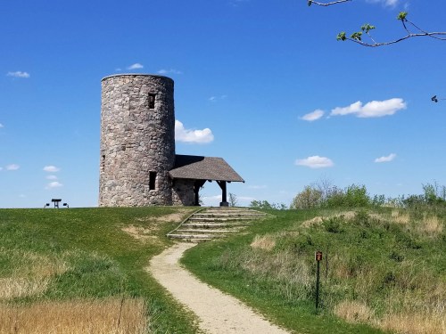 A stone tower with a thatched roof sits atop a grassy hill under a blue sky with scattered clouds.
