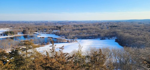 A scenic winter landscape featuring snow-covered fields, a lake, and distant hills under a clear blue sky.