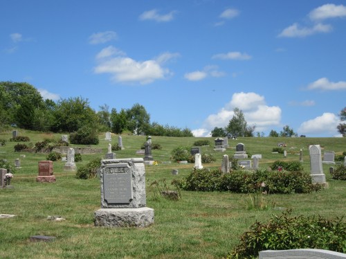 A peaceful cemetery with gravestones on a grassy hill under a blue sky with fluffy clouds.
