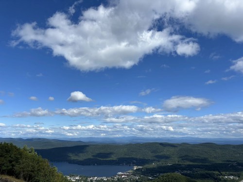 A scenic view of a lake surrounded by green hills under a blue sky with fluffy white clouds.