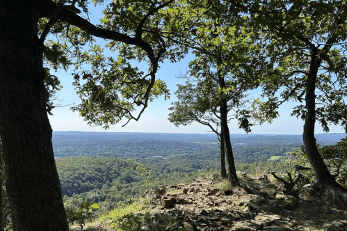 A scenic view from a rocky overlook, framed by trees, showcasing rolling hills and a clear blue sky.
