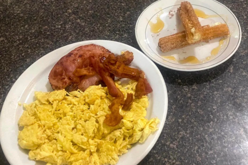 A plate with scrambled eggs, bacon, and ham, alongside a plate of churros drizzled with syrup.
