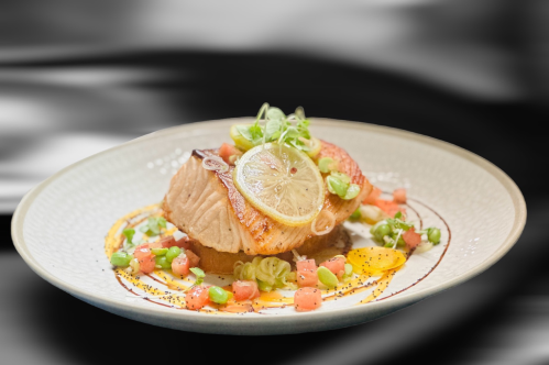 A beautifully plated salmon dish topped with lemon, green onions, and diced vegetables on a decorative plate.